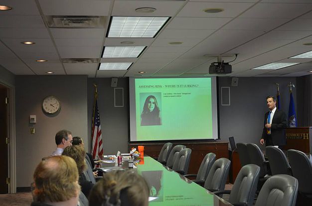 Presenter speaking to seated attendees in a conference room, with a projected slide titled 'Assessing Risk, Where Is It Lurking?