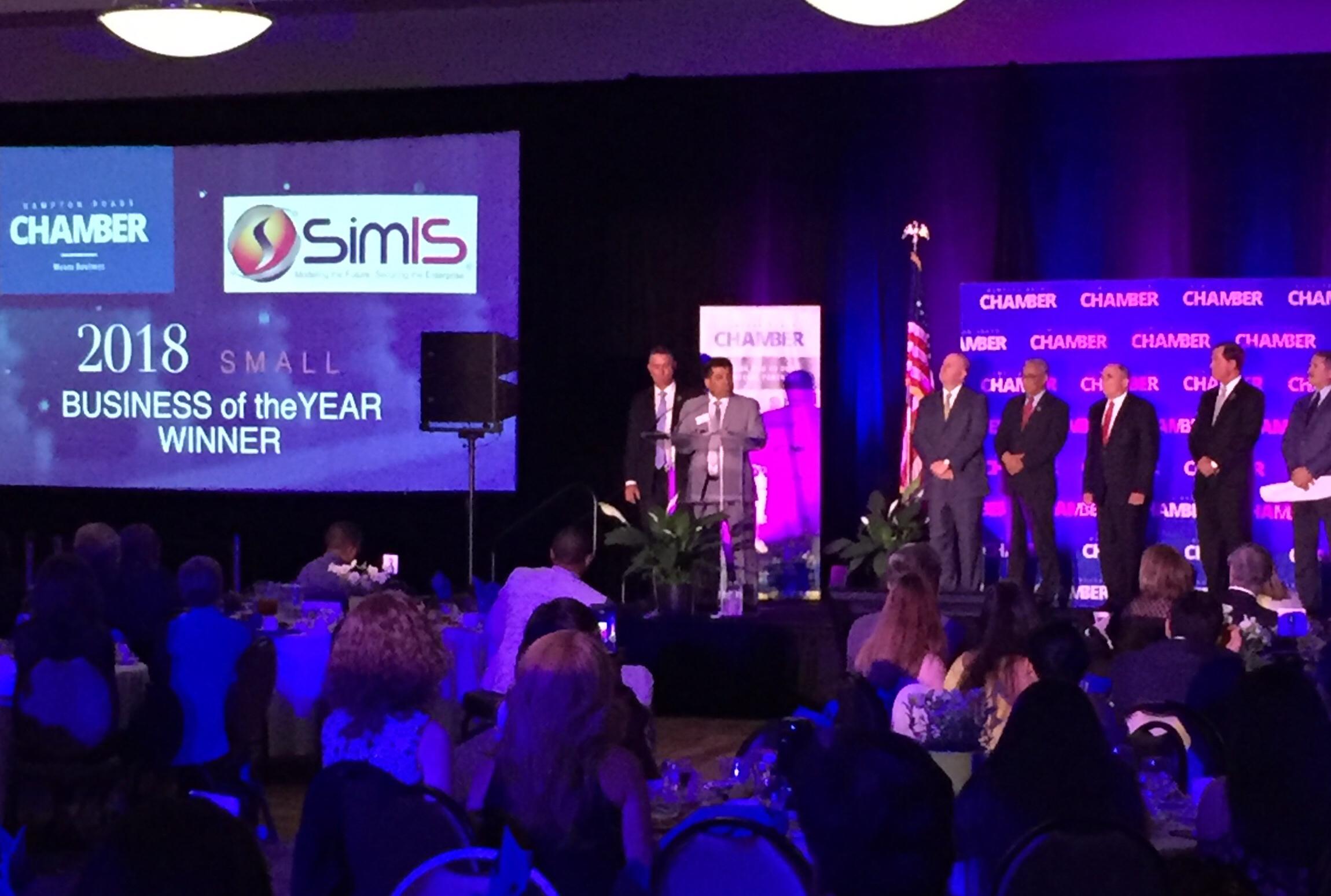 Dr. Johnny Garcia speaking at a podium at the Hampton Roads Chamber awards ceremony, with a screen reading '2018 Small Business of the Year Winner, SimIS.'