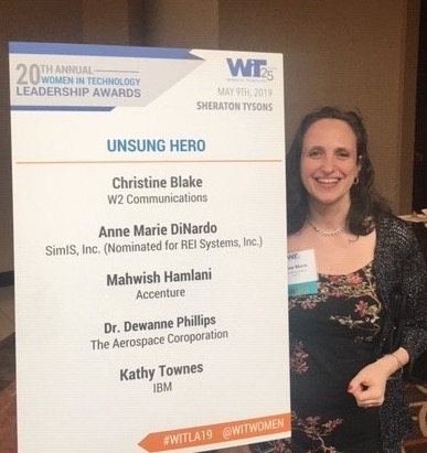 Alt text: "Anne Marie DiNardo posing beside the Women in Technology Leadership Awards Unsung Hero finalist board listing her nomination.